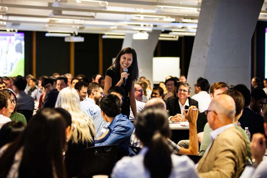 Woman speaking in front of crowd of people sitting at tables.